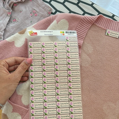 Mano che tiene un foglio di etichette con nome sopra un maglione rosa già etichettato